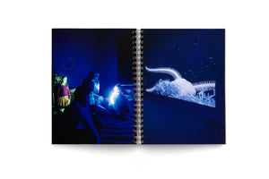 An open, spiral-bound book displays a two-page photograph. The image shows a group of people and children in a dark room with blue lighting, watching a large screen. The screen shows a magnified, white sea creature, likely a basket star, with long, spiny, branching arms against a dark background.