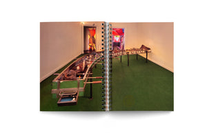A photograph in an open, spiral-bound book shows an art installation in a room with a green floor. The artwork is a very long, custom-made foosball table. Instead of a flat playing field, the center of the table is a trough filled with large, dark, volcanic-like rocks, some of which have small flames lit within them.