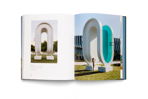 An open art book lies on a white surface, displaying a two-page spread of a large, modern outdoor sculpture. The white sculpture is shaped like an inverted U. The photo on the right page shows a person looking up at the sculpture, revealing its bright turquoise interior that resembles a swimming pool.