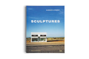 A book titled Sculptures by Elmgreen & Dragset is shown against a white background. The cover features a photograph of the Prada Marfa art installation, a standalone white storefront in a desert landscape under a large, clear blue sky.