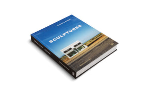 A thick, hardcover art book titled SCULPTURES by Elmgreen & Dragset, set against a plain white background. The cover photo shows the Prada Marfa art installation, a small, white, standalone building in a desert landscape next to a road, beneath a vast blue sky.