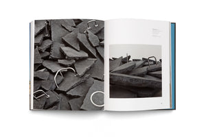 An open book on a white surface displays a two-page spread featuring photographs of a contemporary art installation. The artwork consists of large, broken, charcoal-colored slabs, resembling asphalt, with polished stainless steel fixtures like U-shaped bars embedded within the shattered pieces.
