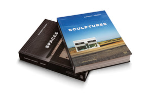 Two hardcover art books by Elmgreen & Dragset are shown against a white background. The top book, titled Sculptures, has a blue cover with a photo of the Prada Marfa art installation. It rests diagonally on the second book, a black volume titled Spaces, which features an architectural image on its cover.