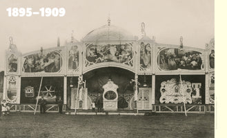A sepia-toned photograph from 1895-1909 of the ornate, two-story facade of a travelling Grand Theater at a fairground. The structure is heavily decorated with Art Nouveau style paintings and carvings. Several men and a few children stand on the dirt ground in front of the large, arched entrance which houses a large fairground organ.
