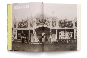 An open book displays a historical black and white photograph from 1895-1909 across a two-page spread. The photo shows the ornate, Art Nouveau-style facade of a large traveling fairground attraction or theater, with several men in suits standing in front of it on a dirt field.