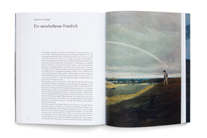 An overhead view of an open book on a white surface. The left page contains German text under the title Ein verschollener Friedrich by Sebastian Schlegel. The right page features a full-page reproduction of a Romantic painting depicting a person standing on a hill overlooking a vast landscape with a large rainbow arcing across the sky.