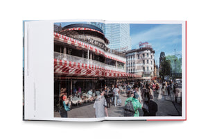 A photograph in an open book showing a composite image that merges past and present views of a busy Berlin street. Translucent figures from an older era walk among modern-day crowds in front of a cafe with red and white striped awnings. Historic buildings and a modern glass skyscraper line the street under a clear blue sky.