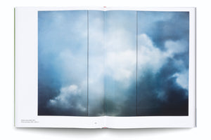 An overhead photo of an open art book on a white background. The two-page spread features a large, atmospheric painting of a turbulent sky, divided into three vertical panels. The painting depicts billowing clouds in shades of deep blue, grey, and white.