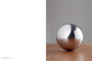 A two-page spread from a book. The left page is white with a caption in the bottom corner. The right page displays a studio photograph of a smooth, matte-finish stainless steel sphere resting on a light-colored wooden surface against a plain grey background.