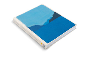 A hardcover book, Ugo Rondinone: Cry Me a River, is displayed at an angle on a white surface. The book features a canvas cover with a minimalist landscape in various shades of blue, depicting water and distant mountains. The off-white spine has the title and other information printed in blue.