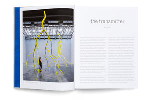 An open art book displaying a two-page spread. The left page features a photograph of a large gallery with towering, bright yellow sculptures that resemble lightning bolts or inverted trees, with a person looking on. The right page contains an essay with the title, the transmitter.