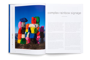 An overhead shot of an open book. The left page features a full-page photograph of the Seven Magic Mountains art installation, showing several totems of brightly colored, stacked boulders in a desert. The right page contains text under the title complex rainbow signage by jarrett earnest.