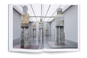 An open book displays a photograph of a modern art gallery exhibition featuring several large, abstract humanoid figures made from stacked, rough-hewn blocks of stone. The sculptures, in shades of grey and tan, stand on flat metal plates on a polished grey floor.