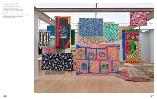 An art gallery installation by Hamid Zénati featuring numerous vibrantly colored, painted textiles hanging from the ceiling. The fabrics, displaying a mix of abstract and geometric patterns, create a dense, layered display in a bright room with light wood floors and several ornate rugs in the background.