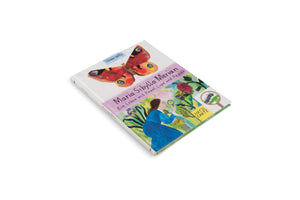A high-angle photo of the book Maria Sibylla Merian: Ein Leben mit Pinsel, Lupe und Papier by Sophie Artz, lying on a white surface. The illustrated cover shows a large peacock butterfly above a drawing of a woman in a blue dress, who is in a garden examining a plant with a magnifying glass.