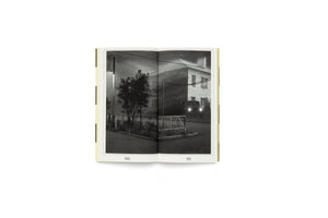 An open book on a white background shows a two-page, black-and-white photograph of a nighttime street scene. On the left page, a street lamp illuminates a fence and tree. On the right page, a van with bright headlights is parked in front of a brick building.