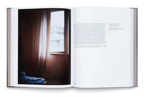 An overhead view of a thick, open art book with a grey cover, lying on a white surface. The left page features a full-bleed, vertical photograph by Nan Goldin titled Hotel Room Zurich, 1988, showing a blurry, sunlit window in a dark room. The right page contains German text about the artwork and artist.