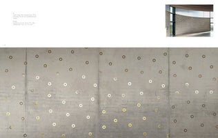 An open art catalog spread. A large close-up photo shows a gray concrete wall decorated with a scattered pattern of small, metallic rings in shades of gold and silver. A smaller inset image in the top right corner shows a wider view of the installation on a curved wall within a modern, sunlit room with large windows.