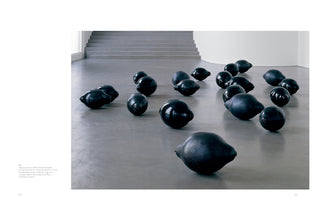 A modern art installation in a minimalist gallery with a grey floor and stairs in the background. The installation consists of numerous large, black, lemon-shaped ceramic sculptures scattered across the floor, with a mix of glossy and matte finishes.