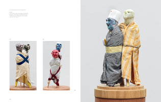An open art book displays several mixed-media sculptures of stylized human figures on wooden bases. The largest image shows two figures tied together; one has a blue face and a white hat, while the other is wrapped in a yellow-and-white striped towel. Two smaller images on the left show other multi-headed figures wrapped in towels and cloth.