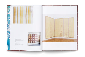 An open hardcover art book on a white surface, displaying a two-page spread with photographs of artworks. The left page shows two images of paintings with vertical yellow and tan stripes alongside text. The right page features a large photo of a room-corner installation of several tall, striped panels.