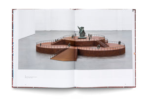 An open art book displays a photograph of a large sculpture in a gallery. The artwork is a multi-tiered, clover-shaped platform made of rusted steel. Small bronze figures of people stand on the different levels, connected by staircases, with a larger bronze monument on the highest tier.