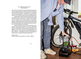 A two-page magazine spread. The left page has German text. The right page shows a person in a striped shirt and patterned pants leaning over a black blender filled with chopped green herbs. Behind them, a black electric bicycle is parked indoors on a wooden floor.