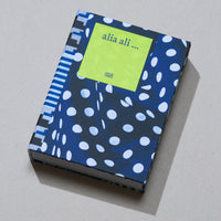 A hardcover book titled alia ali ... lies diagonally on a textured gray surface. The cover is a vibrant blue fabric with a pattern of irregular white polka dots. A contrasting geometric pattern of stripes and blocks in blue, white, and black adorns the spine. A bright lime-green label on the front displays the title and the publisher, Hatje Cantz.