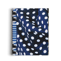 A book stands upright against a white background, casting a soft shadow. Its cover features a dynamic pattern of large, distorted white polka dots on a dark blue background that resembles draped fabric. The spine has a contrasting vertical band with geometric patterns in black, white, and blue.