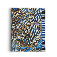 An angled view of a rectangular canvas wrapped in fabric with a traditional batik pattern. The intricate design features circular and abstract shapes in shades of blue, brown, and white with dot work on a black background. A contrasting geometric border with blue, white, and black stripes runs along the right side.