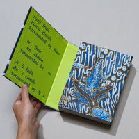 A hand holds open a book with a blue and brown batik-style fabric cover, set against a plain gray background. The inside cover is a vivid lime green, featuring lines of fragmented, poetic text printed in a dark blue font.