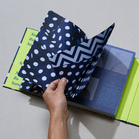 An overhead shot of a person's hand turning a page in a hardcover book. The book has a lime green spine and a dark blue interior. The page being turned is glossy and features a blue and white polka dot pattern on one side and a chevron pattern on the other, revealing a page with blue text and graphics underneath.