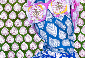 A close-up of a figure draped in several colorful, block-printed fabrics. The figure's head is covered in a bright blue textile with white leaves and a pink wrap featuring two decorative patches that resemble sunglasses with a palm tree pattern. The background is a green fabric with a repeating white and purple leaf-like motif.