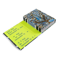 An angled, overhead shot of a book with a blue, brown, and white batik-patterned cover and a vertically striped spine. The book is slightly open, revealing a bright, lime-green inside cover with several lines of black text printed on it, all against a white background.