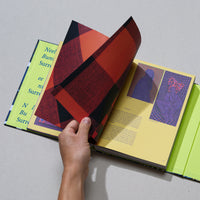 A hand turns a page with a red and black plaid pattern in a colorful art book. The book lies open on a grey surface, revealing yellow pages with text and graphic images, and a bright chartreuse green spine and inside cover.