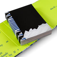 A high-angle, close-up shot of an art book lying in an open, vibrant lime-green case. The book's cover is black with a torn bottom edge that reveals a textured white image underneath. The exposed spine features a bold, geometric pattern in black, white, and blue. Blue text is visible on the inside of the lime green cover.
