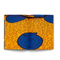 An open book displays a two-page spread of a vibrant African wax print fabric. The pattern features large, circular, shell-like shapes in a deep blue, detailed with black triangles and spirals. These are set against a bright yellow-orange background covered in a dense pattern of small black swirls and lines.