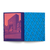 An open notebook on a white surface. The left page features a photograph of an ornate, historic building illuminated at night, rendered in a red and purple duotone. The right page is bright blue with a repeating pattern of green and white stylized leaves.