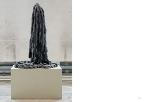 A tall, vertical sculpture made of thick, twisted ropes in shades of dark blue, grey, and brown sits on a white pedestal. The ropes hang densely from the top and coil in a pile at the base, set against a plain concrete wall in a gallery space. The image is on the left side of a page, with a large white space to the right.