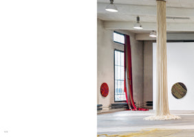 A view of a modern art exhibition in an industrial space with concrete floors. In the center, a thick pillar is covered in cascading off-white ropes that pool on the floor. Two circular, textured artworks hang on the walls: a vibrant red one near a tall window on the left, and an earth-toned one on the right. A long, red and grey textile hangs next to the window.
