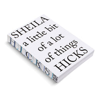 A thick, white paperback book lying on a white surface, viewed from an angle. The cover text in a black serif font reads, SHEILA a little bit of a lot of things HICKS. The fore-edge of the book's pages is decorated with a blue, black, and white abstract pattern.