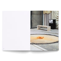 An open book showing a two-page spread. The left page is blank with the number 90 at the bottom. The right page features a photograph of an art installation in a sparse, industrial room with a concrete floor. In the foreground, a circular patch of orange pigment on the floor contains a crumpled orange object. Behind it stands a simple, light-colored stone bench. To the right, a small television on a black pedestal displays a black-and-white image. A large metal door and draped cloths are in the background.