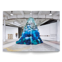 An open book showing a two-page spread of an art gallery with a large, central installation. The piece is a tall, conical pile of soft, rounded cushions in a gradient of blue, from dark teal and navy at the wide base to pale sky blue at the peak. The gallery has an industrial feel with a high ceiling, exposed pipes, and a concrete floor. Other artworks are visible on the white walls in the background.