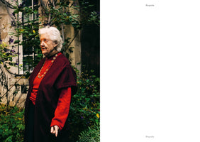 A diptych layout with a color photo on the left and white space on the right. The photo shows an elderly woman with white hair, wearing a red sweater and a dark red shawl, standing in a garden with green vines and purple flowers. The white space has the words Biografie and Biography.