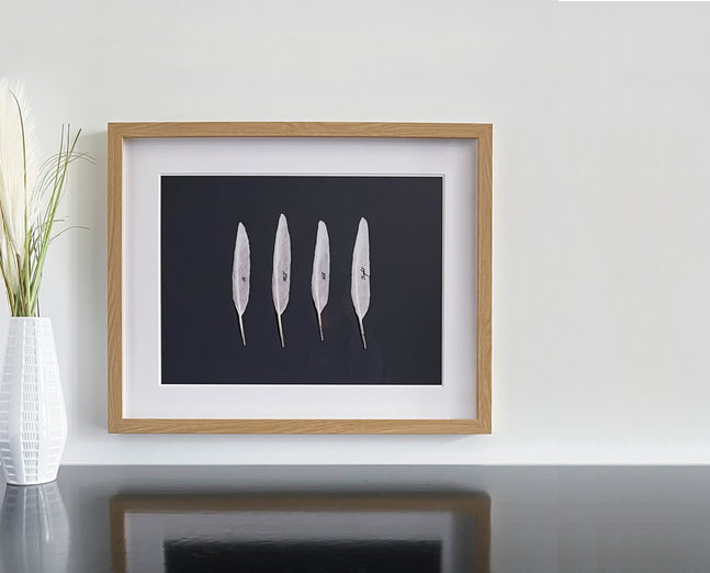 A piece of art in a light wood frame sits on a dark tabletop next to a white vase with tall grass. The art features four white feathers arranged in a row on a black background, with a single word written in script on each feather.
