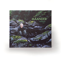 A square hardcover book titled MÄANDER by moki, resting on a white surface. The cover art depicts a person with short blonde hair standing waist-deep in a dark river, surrounded by large, moss-covered rocks in a forest.