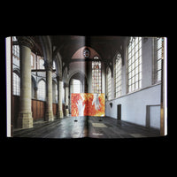 A photograph of an open book on a black background. The two-page spread shows a modern art installation inside a historic church with stone pillars and high, arched windows. The art piece is a large, wide panel displaying two pale hands against a fiery background of orange, yellow, and red.