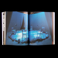 A full-page spread in an open book against a black background, showing a photograph of an art installation. The scene is bathed in blue light from spotlights. Several simple wooden chairs are arranged in a circle, which is outlined by large stones on the floor. Crumpled silver objects hang from the ceiling above the chairs. A caption at the bottom reads, Silver Lake with Dolmen Music.