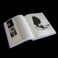 An open book with a purple spine is displayed at an angle on a black background. The pages contain black text and several black-and-white photographs. The right page is dominated by a large photo of a woman with long dark hair lying on the floor, looking upside down at the camera.