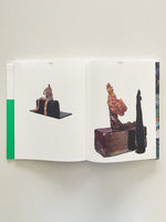 An overhead shot of an open art book on a white surface. The pages display two abstract, lumpy humanoid sculptures against a white background. The sculpture on the left is brown and sits on a black block. The sculpture on the right is multicolored and sits on a larger block next to a tall, thin black object. A bright green strip is visible on the book's left edge.